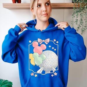 May include: Royal blue hoodie featuring a graphic of Mickey Mouse balloons, a pink bow, and a geometric sphere. The design is framed with a white border and surrounded by small pink flowers. The hoodie has a drawstring hood and a kangaroo pocket.