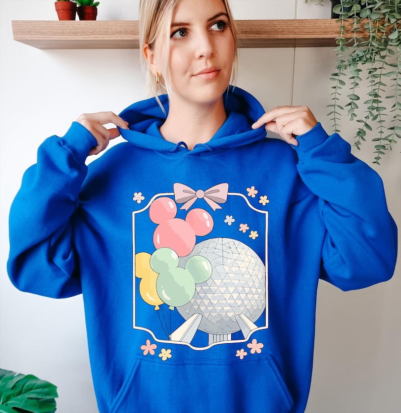 May include: Royal blue hoodie featuring a graphic of Mickey Mouse balloons, a pink bow, and a geometric sphere. The design is framed with a white border and surrounded by small pink flowers. The hoodie has a drawstring hood and a kangaroo pocket.