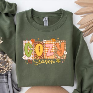 A retro patchwork design spells out “COZY” in colorful letters, each block decorated with autumn patterns like plaid, pumpkins, and florals. Below it, the word “Season” appears in cursive, surrounded by leaves and stars.