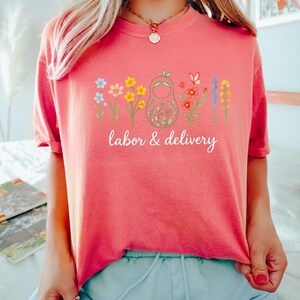 May include: A pink t-shirt with a graphic of a nesting doll surrounded by colorful flowers. The text "labor & delivery" is printed below the graphic.