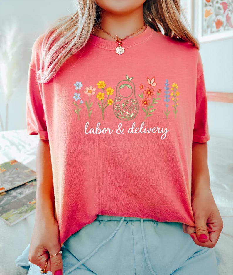 May include: A pink t-shirt with a graphic of a nesting doll surrounded by colorful flowers. The text "labor & delivery" is printed below the graphic.