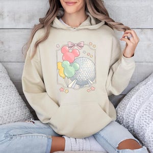 May include: Beige hoodie with a front graphic featuring Mickey Mouse-shaped balloons in pink, yellow, and green, along with a silver sphere and a pink bow. The design is framed with a light gray border and small pink flowers.