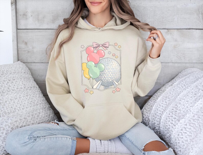 May include: Beige hoodie with a front graphic featuring Mickey Mouse-shaped balloons in pink, yellow, and green, along with a silver sphere and a pink bow. The design is framed with a light gray border and small pink flowers.