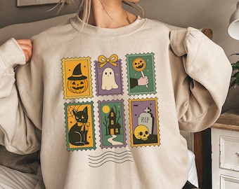 May include: Beige sweatshirt featuring a colorful Halloween-themed stamp design. The stamps depict a jack-o'-lantern, ghost, lollipop, black cat, haunted house, and skull with a tombstone. The design is outlined in black and has a vintage aesthetic.