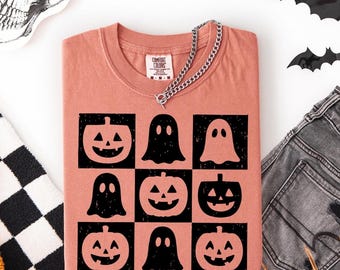 May include: A coral-colored t-shirt featuring a grid pattern of black jack-o'-lanterns and ghosts. The shirt has a crew neck and short sleeves. A silver chain necklace is draped across the shirt. Halloween-themed apparel.