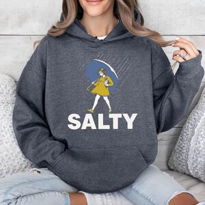 K&ouml;nnte beinhalten: Dunkelgrauer Kapuzenpullover mit Frontaufdruck. Der Aufdruck zeigt eine Cartoonfigur in einem gelben Kleid und mit blauem Regenschirm, darunter das Wort "SALTY" in wei&szlig;en Blockbuchstaben. Der Hoodie hat eine Fronttasche und eine Kapuze mit Kordelzug.