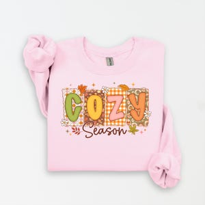 K&ouml;nnte beinhalten: Ein hellrosa Sweatshirt mit dem Schriftzug "COZY Season" in einem farbenfrohen, herbstlichen Design. Die Buchstaben sind in verschiedenen Mustern und Farben gehalten, darunter Gr&uuml;n, Orange und Rosa, mit floralen und Blatt-Akzenten. Das Sweatshirt hat lange &Auml;rmel.
