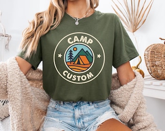 May include: Olive green t-shirt with a circular graphic that reads "CAMP CUSTOM." The graphic features a mountain scene with a tent, sun, and trees. The shirt is short-sleeved and has a relaxed fit.