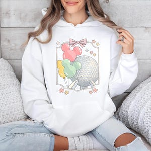 May include: White hoodie featuring a colorful graphic of Mickey Mouse-shaped balloons, a pink bow, and a geometric sphere. The design includes yellow, pink, and green balloons. The hoodie is a comfortable, casual style.