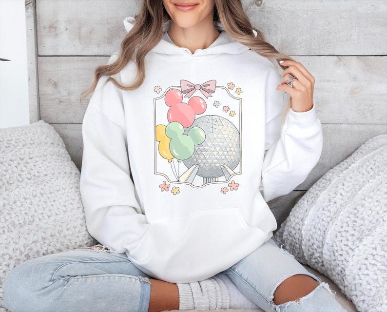 May include: White hoodie featuring a colorful graphic of Mickey Mouse-shaped balloons, a pink bow, and a geometric sphere. The design includes yellow, pink, and green balloons. The hoodie is a comfortable, casual style.