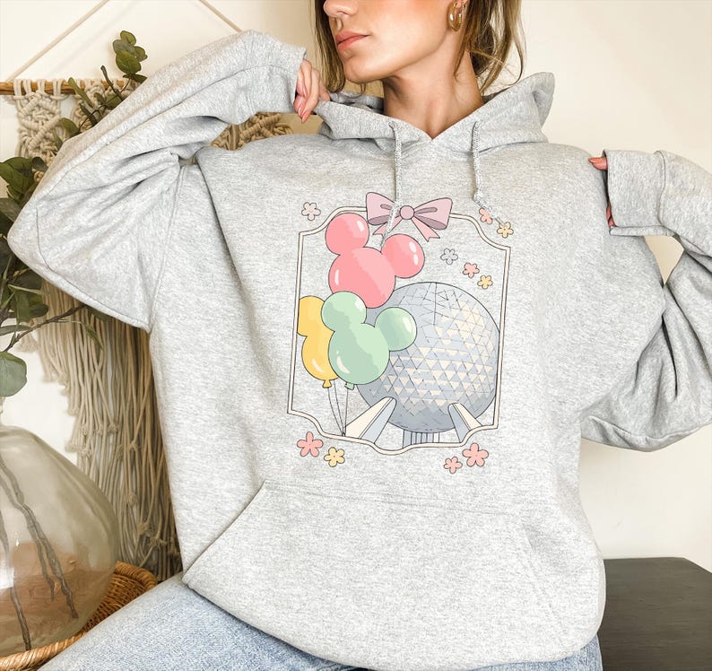 May include: A light gray hoodie featuring a colorful graphic. The graphic includes Mickey Mouse-shaped balloons in pink, yellow, and green, a bow, and a depiction of a geodesic sphere. Floral accents surround the central design.