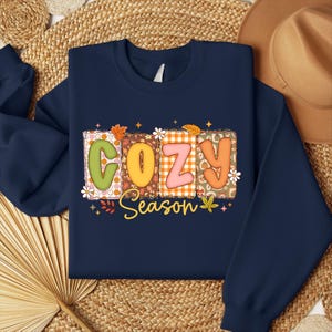 K&ouml;nnte beinhalten: Marineblaues Sweatshirt mit dem Schriftzug "COZY Season" in einem farbenfrohen, herbstlichen Design. Die Buchstaben sind mit Mustern gef&uuml;llt und mit Bl&auml;ttern und Blumen verziert. Ein gem&uuml;tliches Herbst-Themen-Kleidungsst&uuml;ck.
