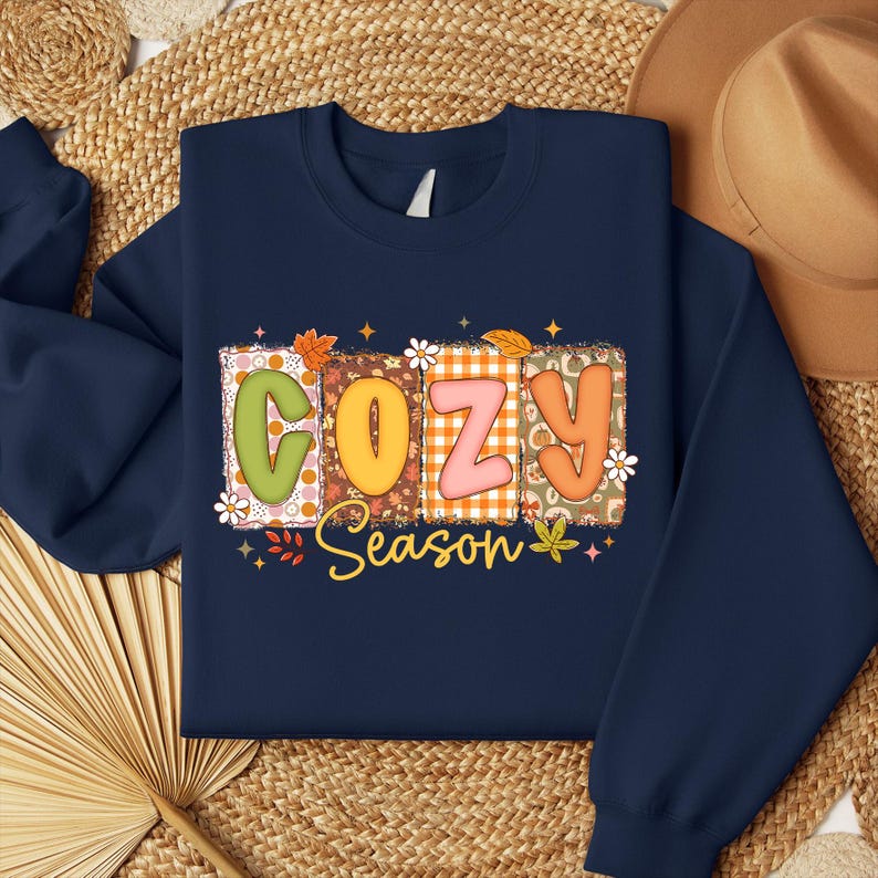 K&ouml;nnte beinhalten: Marineblaues Sweatshirt mit dem Schriftzug "COZY Season" in einem farbenfrohen, herbstlichen Design. Die Buchstaben sind mit Mustern gef&uuml;llt und mit Bl&auml;ttern und Blumen verziert. Ein gem&uuml;tliches Herbst-Themen-Kleidungsst&uuml;ck.