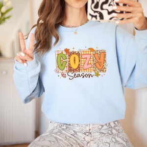 K&ouml;nnte beinhalten: Hellblaues Sweatshirt mit dem Schriftzug "COZY Season" in einem farbenfrohen Retro-Design. Die Buchstaben sind gr&uuml;n, orange und rosa, mit floralen und Blatt-Akzenten. Das Sweatshirt hat lange &Auml;rmel.