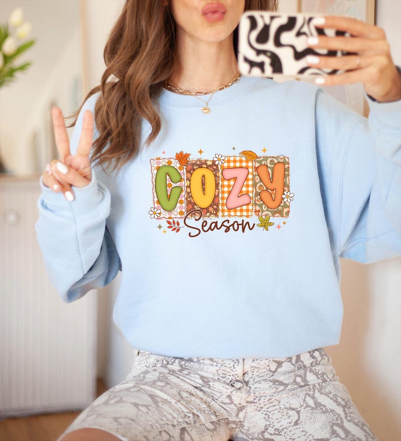 K&ouml;nnte beinhalten: Hellblaues Sweatshirt mit dem Schriftzug "COZY Season" in einem farbenfrohen Retro-Design. Die Buchstaben sind gr&uuml;n, orange und rosa, mit floralen und Blatt-Akzenten. Das Sweatshirt hat lange &Auml;rmel.