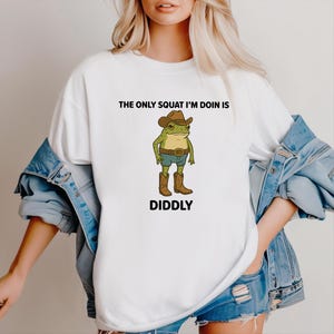 May include: White t-shirt featuring a cartoon frog wearing a cowboy hat, boots, and shorts. The text "THE ONLY SQUAT I'M DOIN IS DIDDLY" is printed above and below the frog illustration. The shirt is a casual, unisex style.