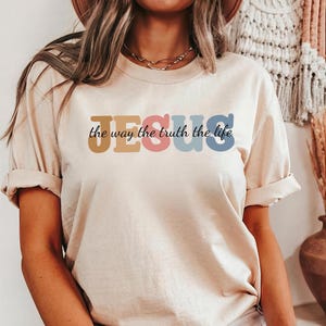 Könnte beinhalten: Beiges T-Shirt mit hochgekrempelten Ärmeln. Auf der Vorderseite steht das Wort "JESUS" in Braun, Rosa, Blau und Blau, darunter der Schriftzug "the way the truth the life" in schwarzer Schrift. Lässiger Stil.