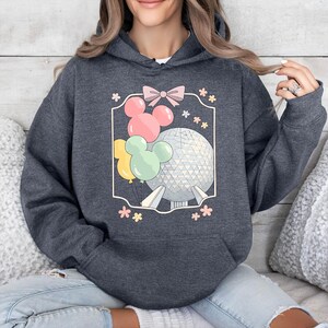 May include: Dark gray hoodie featuring a graphic of a sphere, Mickey Mouse balloons, and flowers. The design is enclosed in a white frame with a pink bow at the top. The hoodie has a drawstring hood and a front pocket.