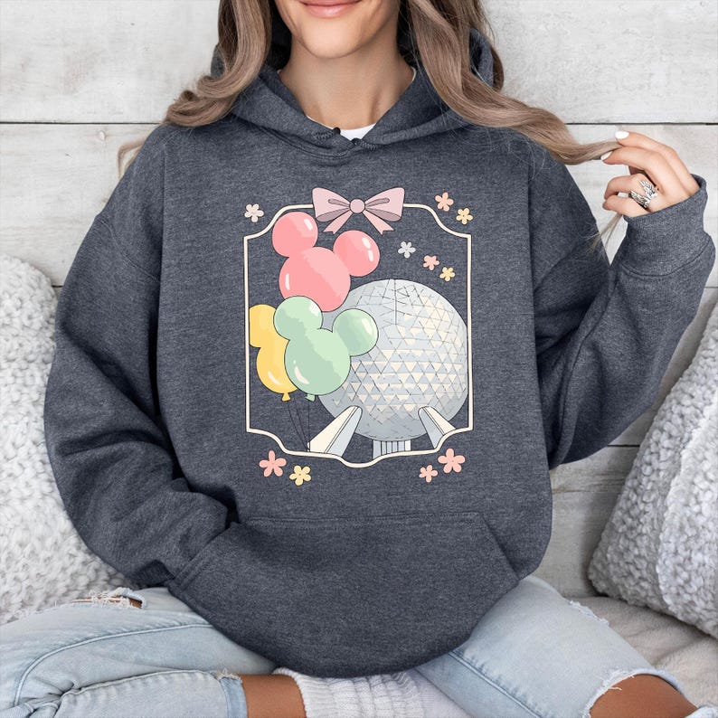 May include: Dark gray hoodie featuring a graphic of a sphere, Mickey Mouse balloons, and flowers. The design is enclosed in a white frame with a pink bow at the top. The hoodie has a drawstring hood and a front pocket.