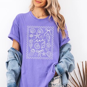 Könnte beinhalten: Ein lila T-Shirt mit einem weißen rechteckigen Design mit Illustrationen zum Thema Meer, darunter Muscheln, Seesterne und Seepferdchen. Das T-Shirt hat hochgekrempelte Ärmel und wird mit einer Jeansjacke getragen.