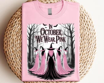 May include: Pink t-shirt with the text "In October We Wear Pink". The graphic features a group of witches in pink dresses and black hats standing in front of a forest backdrop. Bats are flying above the text.