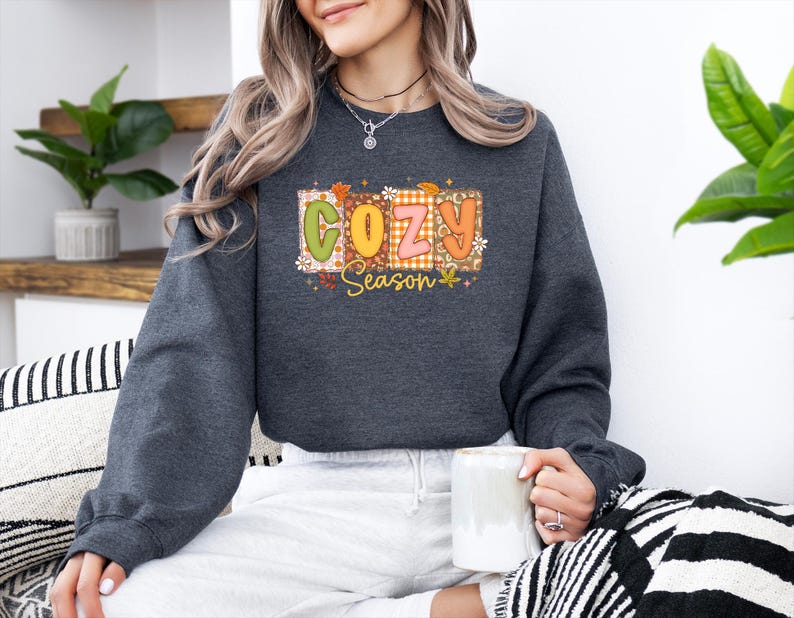 K&ouml;nnte beinhalten: Dunkelgraues Sweatshirt mit dem Schriftzug "COZY Season" in einem farbenfrohen, herbstlichen Design. Der Text befindet sich in einem rechteckigen Rahmen mit Herbstbl&auml;ttern und einem Karomuster. Das Sweatshirt hat lange &Auml;rmel.