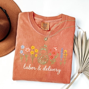 May include: A peach-colored t-shirt with a floral design and the text "labor & delivery".