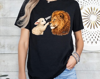 Könnte beinhalten: Ein schwarzes Kurzarm-T-Shirt mit einer Grafik eines weißen Lamms, das die Nase eines braunen Löwen berührt. Der Text BOOP! steht über dem Lamm und der Text LUKE 10:19 ist in kleinen Buchstaben in der Nähe der Löwenmähne aufgedruckt.