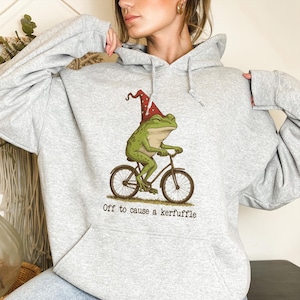 May include: A light gray hoodie featuring a whimsical illustration of a green frog riding a bicycle and wearing a red and white hat. The text "Off to cause a kerfuffle" is printed below the image.