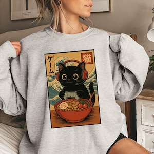 May include: A light gray sweatshirt featuring a graphic of a black cat wearing headphones and holding a game controller. The cat is in front of a bowl of ramen with Japanese text. The background has a wave design.
