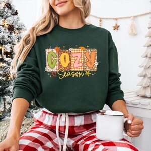 K&ouml;nnte beinhalten: Dunkelgr&uuml;nes Sweatshirt mit dem Schriftzug "COZY Season" in einem farbenfrohen, herbstlichen Design. Die Buchstaben sind im Patchwork-Stil mit floralen und Blatt-Akzenten gehalten. Das Sweatshirt hat lange &Auml;rmel.