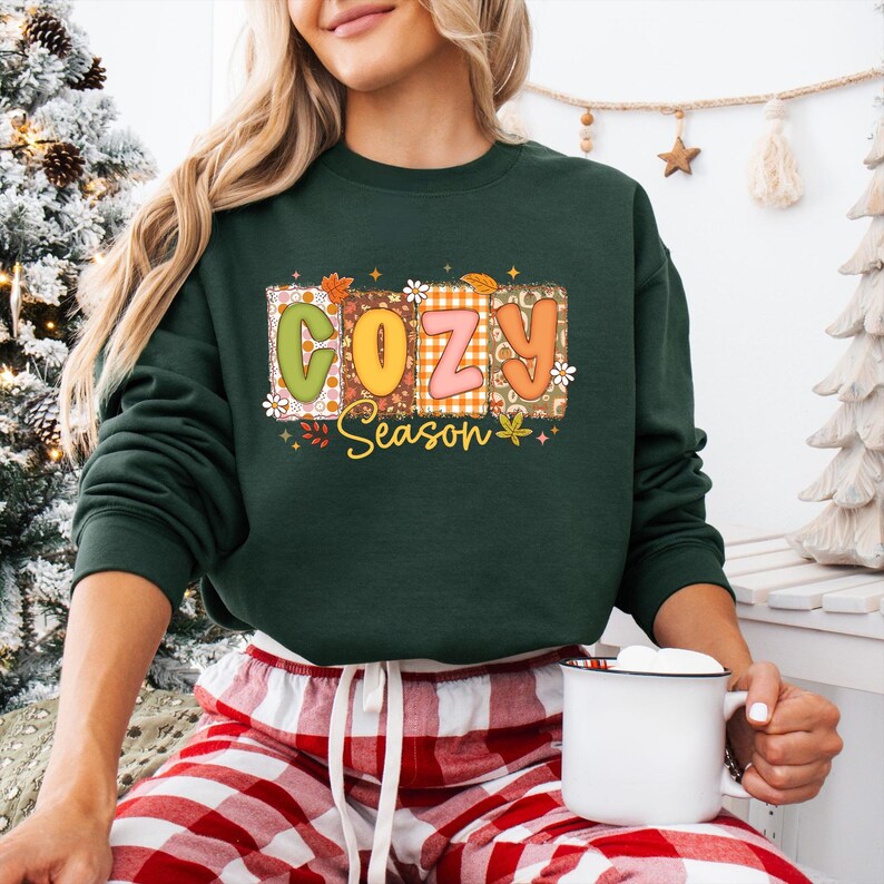 K&ouml;nnte beinhalten: Dunkelgr&uuml;nes Sweatshirt mit dem Schriftzug "COZY Season" in einem farbenfrohen, herbstlichen Design. Die Buchstaben sind im Patchwork-Stil mit floralen und Blatt-Akzenten gehalten. Das Sweatshirt hat lange &Auml;rmel.
