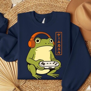 May include: Navy blue sweatshirt featuring a cartoon frog wearing orange headphones and holding a video game controller. The frog is green and yellow. The sweatshirt has Japanese text in an orange rectangle.