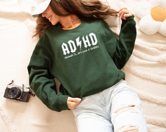 May include: A dark green sweatshirt with the text "ADHD" in a white, stylized font with a lightning bolt graphic. Below the main text, the phrase "HIGHWAY TO...HEY LOOK A SQUIRREL" is printed in white. The sweatshirt is a crew neck style.