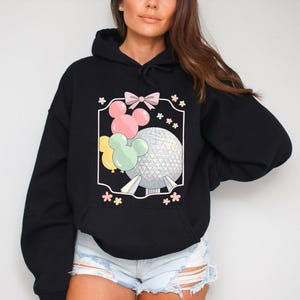 A hooded sweatshirt graphic showing the Spaceship Earth sphere and balloons shaped like Mickey Mouse. Delicate flowers and a ribbon bow surround the main elements. The image evokes feelings of a Disney family holiday and vintage style fashion.