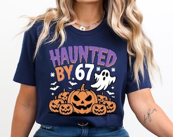 May include: Navy blue t-shirt with a Halloween-themed graphic. The design features the text "HAUNTED BY 67" in purple and orange, along with a ghost, pumpkins, bats, and spiders. The shirt is a short-sleeved crew neck.