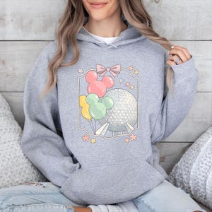 May include: A light gray hooded sweatshirt featuring a colorful graphic. The graphic includes Mickey Mouse-shaped balloons in pink, yellow, and green, along with a geometric sphere and a pink bow. Floral accents surround the design.