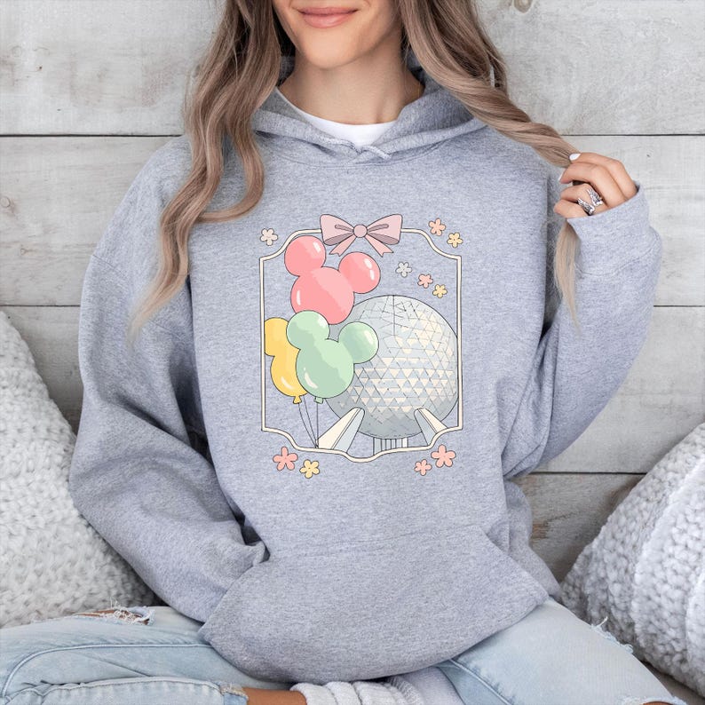 May include: A light gray hooded sweatshirt featuring a colorful graphic. The graphic includes Mickey Mouse-shaped balloons in pink, yellow, and green, along with a geometric sphere and a pink bow. Floral accents surround the design.