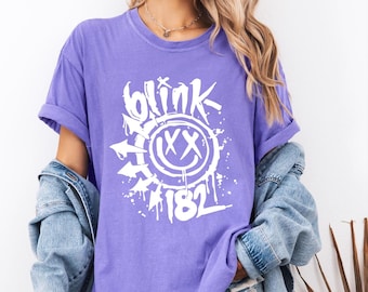May include: A purple t-shirt with rolled-up sleeves featuring a white graphic design. The design includes the word "blink" in a graffiti-style font, a smiley face with "XX" eyes, and the number "182".