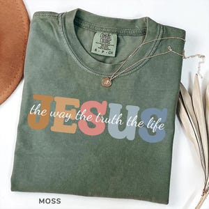 Könnte beinhalten: Moosgrünes T-Shirt mit dem Wort "JESUS" in Retro-Lettern und dem Satz "the way the truth the life". Eine goldene Halskette mit Anhänger liegt auf dem Shirt. Das Shirt ist mit "Comfort Colors" gekennzeichnet.