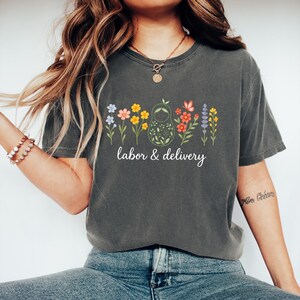 May include: A dark gray t-shirt with a floral design and the text "labor & delivery". The design features a nesting doll with flowers around it.