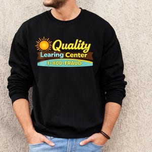 May include: Black sweatshirt with a graphic that reads "Quality Learing Center" in yellow script, with a sun graphic and the phone number "1-800-FRAUD" in white. The sweatshirt is worn by a person.