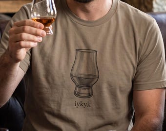 Könnte beinhalten: Ein beiges T-Shirt mit Rundhalsausschnitt und einer schwarzen Strichzeichnung eines tulpenförmigen Whiskyglases, unter der der Text iykyk gedruckt ist. Das T-Shirt wird von einer Person getragen, die ein passendes Glas mit bernsteinfarbener Flüssigkeit hält.
