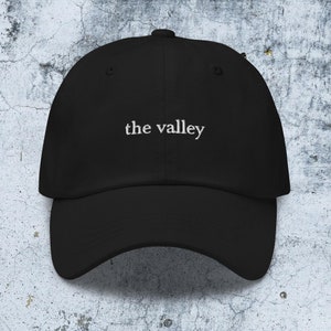 May include: A black baseball cap featuring the text "the valley" embroidered in white thread across the front center. The cap has a structured crown, a curved brim, and a button at the top, presenting a clean and simple design for headwear.