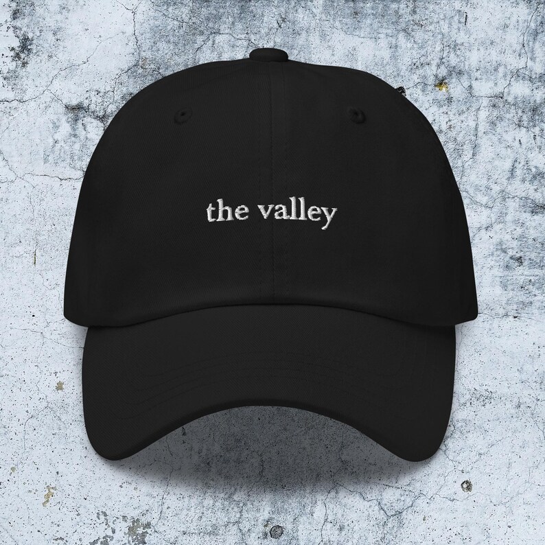 May include: A black baseball cap featuring the text "the valley" embroidered in white thread across the front center. The cap has a structured crown, a curved brim, and a button at the top, presenting a clean and simple design for headwear.