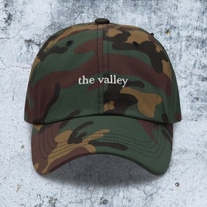 May include: A camouflage baseball cap featuring a green, brown, and black pattern. The front of the cap displays the text 'the valley' embroidered in white thread. The cap has a curved brim and a button at the top center of the crown.