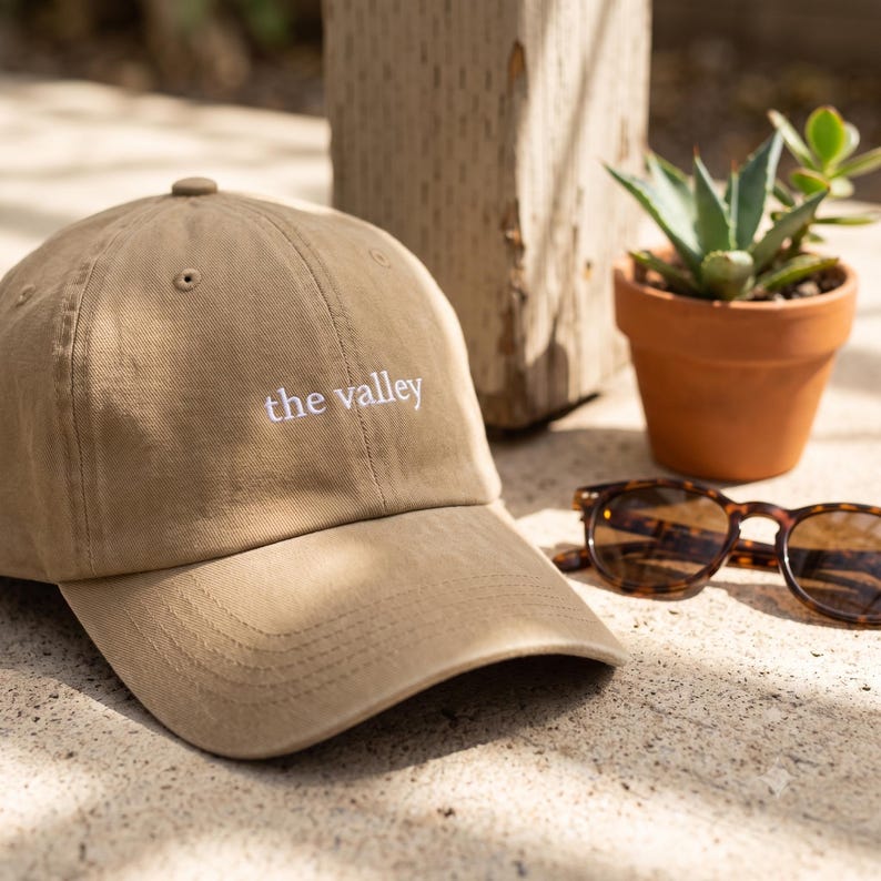 May include: A tan baseball cap featuring the text "the valley" embroidered in white thread on the front panel. The cap has a curved brim, a button on the top, and stitched eyelets for ventilation, presenting a classic six-panel construction.