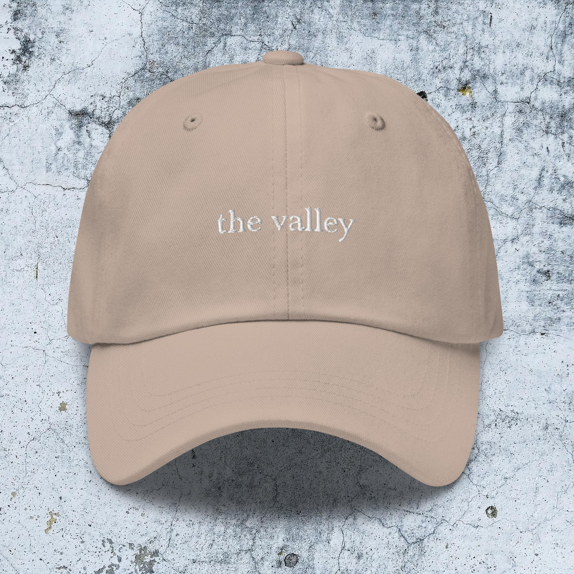 May include: A beige baseball cap featuring the text "the valley" embroidered in white thread across the front center. The cap has a structured crown, a curved brim, and stitched eyelets on the top panels for ventilation and a classic design.