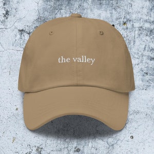 May include: A tan baseball cap featuring the text "the valley" embroidered in white thread across the front center. The cap has a structured crown, a curved brim, and a button at the top, showcasing a clean and simple design with visible stitching.