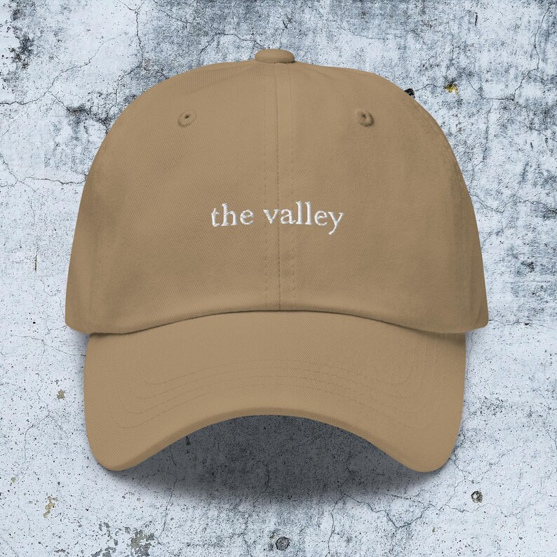 May include: A tan baseball cap featuring the text "the valley" embroidered in white thread across the front center. The cap has a structured crown, a curved brim, and a button at the top, showcasing a clean and simple design with visible stitching.